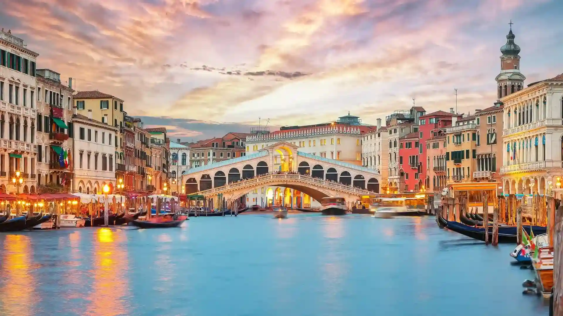 View of the Grand Canal in Venice, Italy, featuring the Rialto Bridge at sunset, surrounded by historic buildings and gondolas on the water