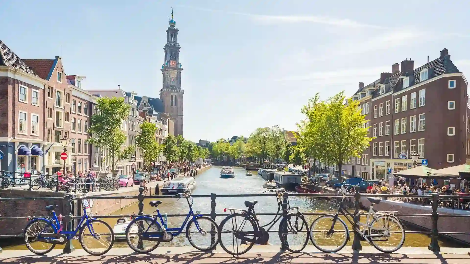 A sunny Amsterdam scene featuring bicycles on a bridge, a canal with boats, and the iconic Westerkerk tower in the background.
